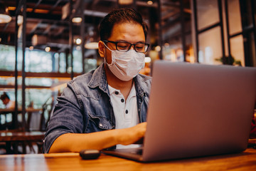 Asian young man indoors in cafe. Lifestyle concept photo with copy space. Picture with handsome guy who using protective face mask. Portrait with gray laptop