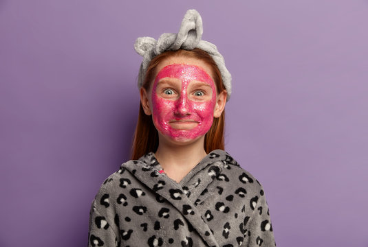 Portrait Of Happy Small European Child Imitates Mother, Applies Pink Beauty Face Mask, Wears Headband And Soft Robe, Enjoys Cosmetic Treatments, Poses Against Purple Background. Useful Cosmetics