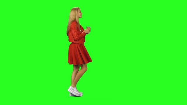 Blonde girl calmly walking and drinking coffee on green screen background. Chroma key, 4k shot. Profile view.