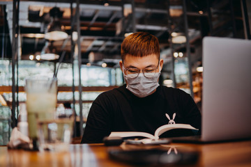 Asian young man indoors in cafe. Lifestyle concept photo with copy space. Picture with handsome guy who using protective face mask and reading book. Portrait with gray laptop