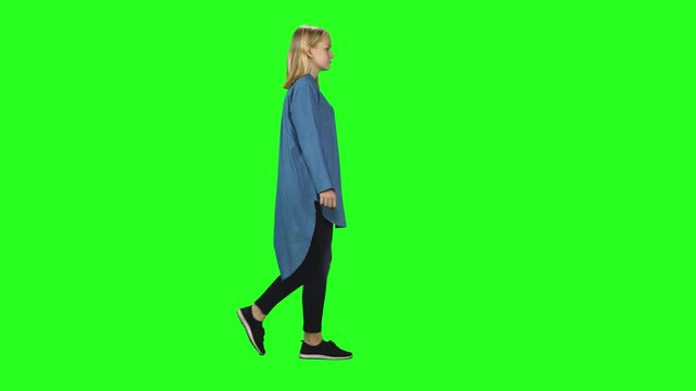 Blonde teenager girl calmly walking on green screen background. Chroma key, 4k shot. Profile view.