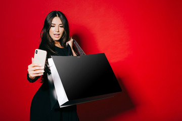 Beautiful stylish young woman in cocktail dress is holding shopping bags, making selfie on smartphone on red background.