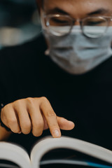 Asian young man indoors in cafe. Lifestyle concept photo with copy space. Picture with handsome guy who using protective face mask and reading book. Portrait with gray laptop