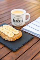 Crispy cereal toast with peanut butter, bananas, chocolate and a caffe latte on the wooden breakfast table.