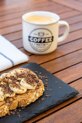 Crispy cereal toast with peanut butter, bananas, chocolate and a caffe latte on the wooden breakfast table.