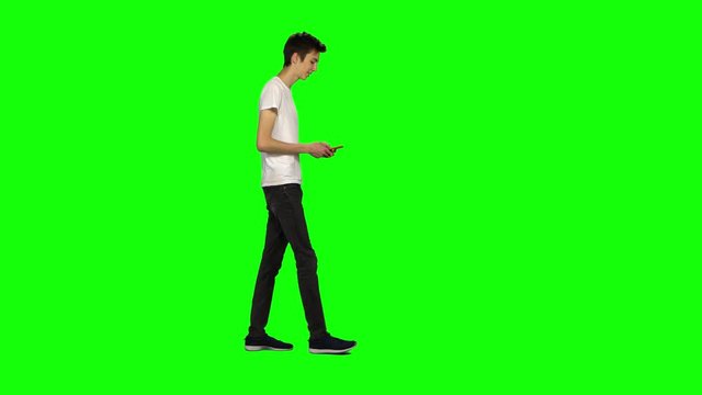 Tall skinny teen guy calmly walking and texting message vie his mobile phone on green screen. Chroma key. Profile view.