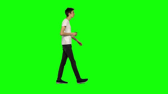Tall skinny teen guy calmly walking and drinking coffee on green screen background. Chroma key, 4k shot. Profile view.
