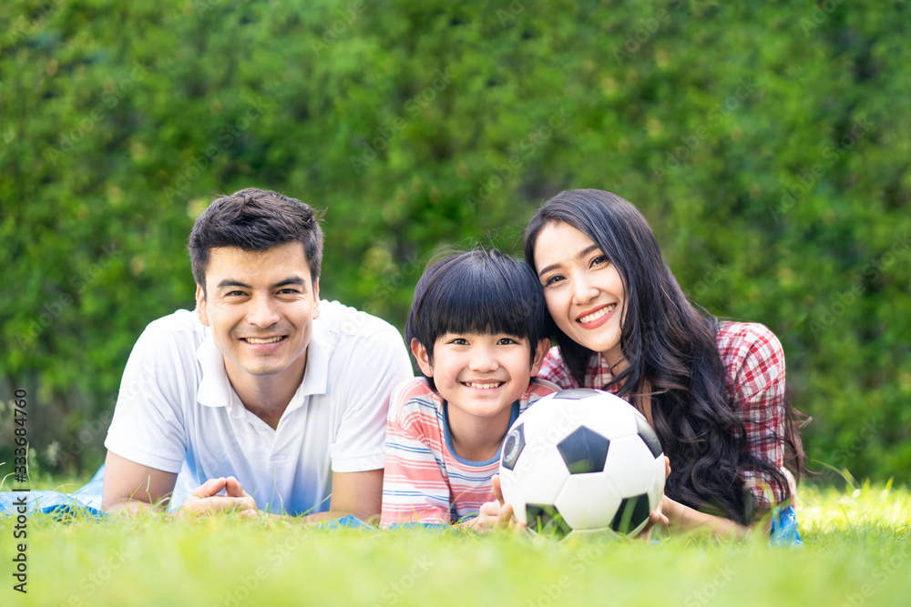 Wall mural Asian lovely young family are in garden. They lay down on grass and smile with happiness moment. Mother and son hold ball. They love to have leisure and be together. Outdoor lifestyle family concept.