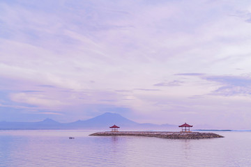 Sunrise on Sanur beach in Bali. Agung volcano in the dawn rays of the pink sun. Traditional gazebos on an artificial island in the ocean. Calm surface of the water.