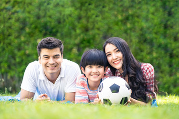 Asian lovely young family are in garden. They lay down on grass and smile with happiness moment. Mother and son hold ball. They love to have leisure and be together. Outdoor lifestyle family concept.