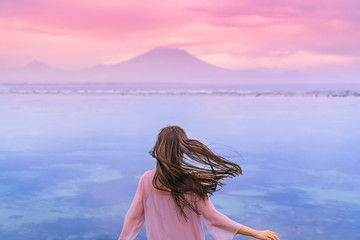 Adventurous young woman in a pink dress blown by the wind looks at the ocean and mountains in the...