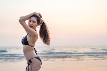 Sexy Asian beautiful smile woman in black bikini holding hair portrait on beach in evening sunset twilight sky. Small wave in sea on background during summer season. Holiday and vacation on the beach.