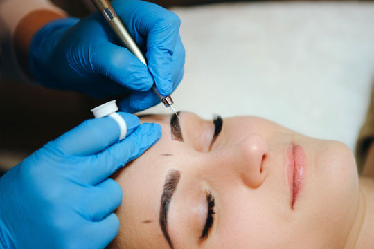 Permanent make-up for eyebrows of beautiful brunette woman in beauty salon. Closeup beautician in gloves doing microblading for eyebrow.