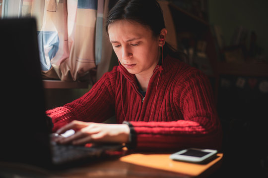 Young caucasian girl in red sweater with confused look on her face looking at computer while studying online at home with pen in hand near the window