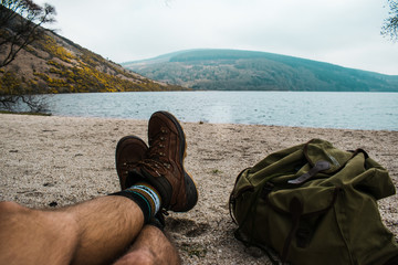 Piernas de un viajero con botas de montaña descansando junto a un lago en Irlanda