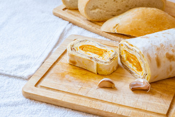 Sliced roll of Armenian pita bread with carrot filling on a wooden board. Sliced white bread in the background. On the board are cloves of garlic. Tasty and healthy homemade treat.