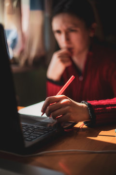 Young caucasian girl in red sweater with confused look on her face looking at computer while studying online at home with pen in hand near the window