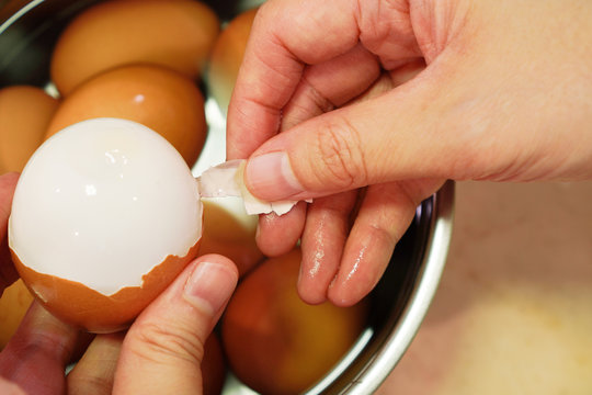 hands hold chicken boiled eggs and peel its shell off with group of eggs in warm water in stainless pot in background ,selective focus on broken shell and tip of right hand thumb