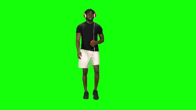 Afro american man is walking in big red headphones, chooses music on mobile phone and moves to rhythm at Green Screen