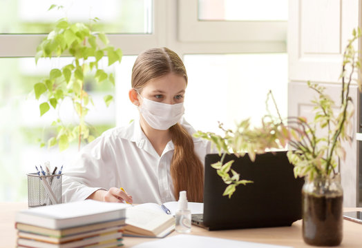 The girl studies at home in a protective face mask during Quarantine CoVid-19. Distance learning online with a laptop. The child is doing homework for school. The schoolgirl stayed at home.
