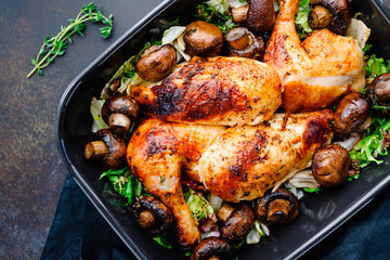 Top view of roasted chicken legs and breasts with fresh salad and mushrooms in black dish.
