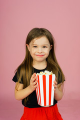 Happy cute little girl holding popcorn in her hands and looking at the camera on a pink background. The concept of watching a movie in a movie theater or at home. Copyspace