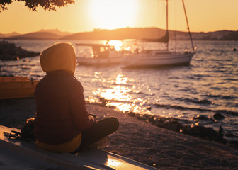 A girl in a sweatshirt looks at the sunset on the sea and at the boats on the shore in the sunshine