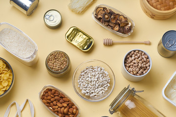 Creative flatlay with pantry staples. Glass jars with pasta, beans and chickpeas, canned goods, nuts and dried mushrooms. Top view pattern with basic products