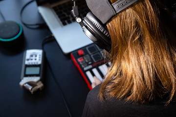 Close up of a female music producer with headphones in her home studio, desk with digital recorder, Smart Home Assistant, notebook and a Midi keyboard. 