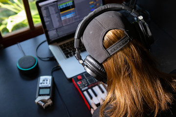 Close up of a female music producer with headphones in her home studio, desk with digital recorder, Smart Home Assistant, notebook and a Midi keyboard. Window with nature in the background.