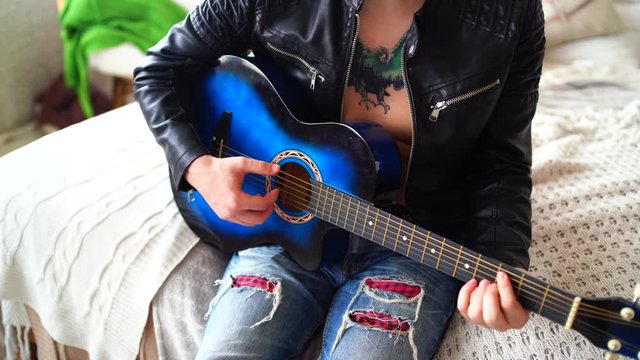 A young man with a tattoo on his chest plays the guitar in the bedroom. Hipster learn to play the guitar. Close-up of a man's hand playing a guitar string. Concept: rock musician