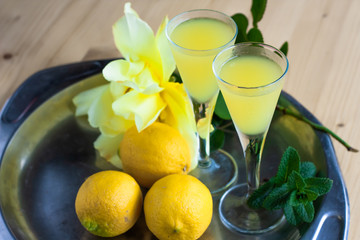 Limoncello in elegant liquor glasses with lemons, mint and yellow rose on steel plate on wooden table. Sicilian lemon liquor aromatic and refreshing. Top view