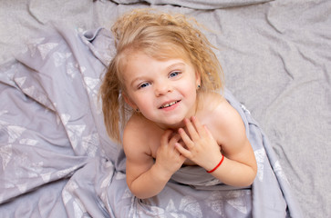 Portrait of a blue-eyed little girl with blond curly hair waking up in the morning on a gray bed laughing merrily.