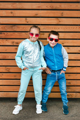 Fashion kids in the city stands on a wooden wall. Trendy boy and girl in sunglasses standing on the street