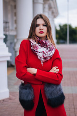 A beautiful, young, fashionable woman in a red sweater is standing on a city street against a building with columns in cool, cloudy, autumn weather.