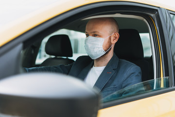 Bald man taxi driver in medical face mask inside yellow car looks at camera, concept of coronavirus quarantine