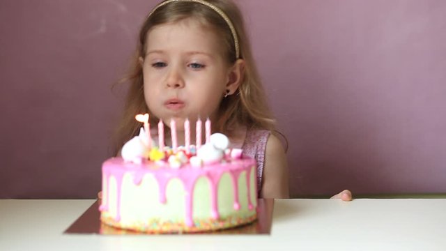 A little girl with blond hair makes a wish and blows out the candles on a pink cake. The day of the birth. Holiday