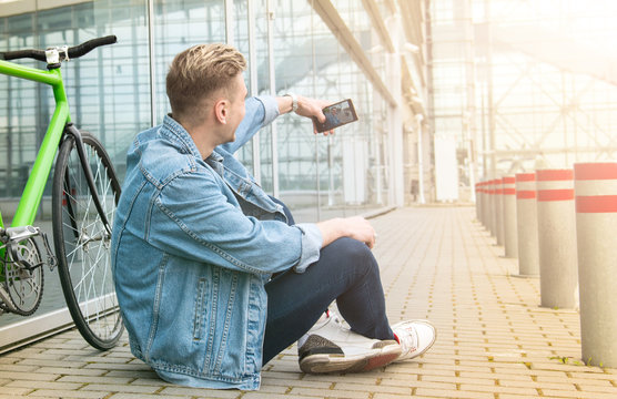 The guy in a blue denim jacket sitting on the street . a young man near green bicycle. Smiling student with looking at phone. A young boy makes selfie.