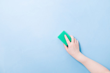 hand without a glove holds a green sponge for washing dishes and cleaning, blue background copy...
