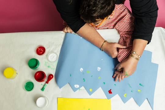 profesora educaci&oacute;n especial y alumno con bata est&aacute;n pintando con las manos una corona de cumplea&ntilde;os y una cartulina amarilla con pinturas acrilicas sobre la mesa. terapia de manualidades pl&aacute;sticas.