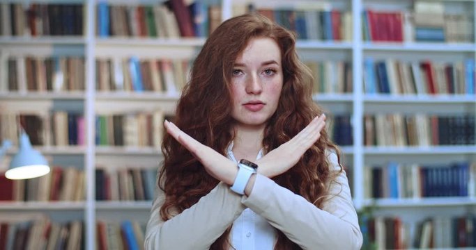 Portrait of beautiful scared young red head girl crossing her hends and saying no in library.Bookcase in background.