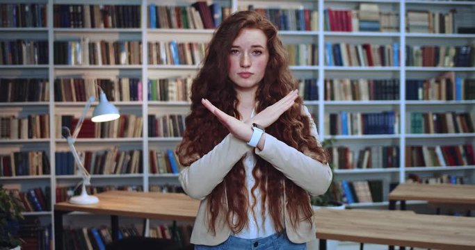 Portrait of pretty sad young red head girl with smart watch crossing her hends and saying no in library. Bookcase in background.