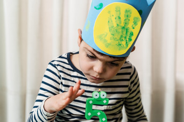 niño de 5 años celebra su cumpleaños durante el confinamiento coronavirus covid19, juega con el pastel y la vela, lleva corona hecha a mano, camiseta a rayas y chandal azul oscuro. es  caucásico.