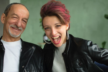 Indoor family portrait of adult daughter and senior father in loft room with houseplants. Crazy man and girl with pink hair are wearing black leather jackets in punk style and smiling