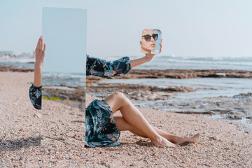 illusion portrait Stylish girl in sun glasses and dress sit on sea beach holding mirrors with reflection of sand and stones. New vision of fashion projects concept. Digital narcissism - obrazy, fototapety, plakaty
