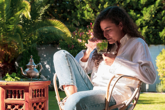 Arabian young woman with ethnic features smelling peppermint arabic tea at her garden. Moroccan female preparing traditional arab tea at home. Arabian culture and traditions. Muslim lifestyle.