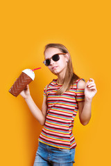 Cute caucasian young girl in t-shirt and sunglasses drinking milk cocktail on yellow studio background