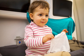 happy little cute baby boy sitting in the baby bassinet of the airplane, smiling and holding a wet, hot towel for washing hands and face after sleeping on a long night flight.