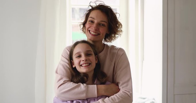 Happy affectionate young mom hugging teen daughter looking at camera standing at home. Smiling adult parent single mother embracing cute teenage adolescent girl posing together indoor. Family portrait