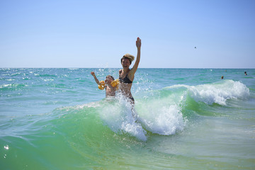 Summer happy family of six years blonde child playing and jumping water waves embracing woman mother in sea shore beach